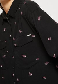 Svart skjorta med en liten paisleymönster i lila och vitt, med två bröstfickor och knäppning framtill.