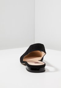Zapato plano de ante negro con laterales recortados, puntera redonda y un tacón negro liso. El interior es de color claro con un pequeño detalle del logo.