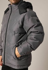 Grijze pufferjas met een ritssluiting aan de voorkant, zijzakken en een contrasterende zwarte capuchon, gemaakt van een getextureerde, waterbestendige stof.