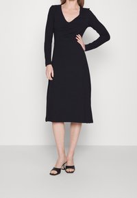 Robe noire à manches longues avec un décolleté en V et des détails froncés au niveau de la poitrine, offrant une coupe décontractée et arrivant au mi-mollet. Tissu en tricot lisse.