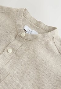 Maglia henley beige texturizzata in tessuto intrecciato. Presenta un colletto alla coreana, due bottoni e una tasca sul petto, con un motivo a quadretti sottile.