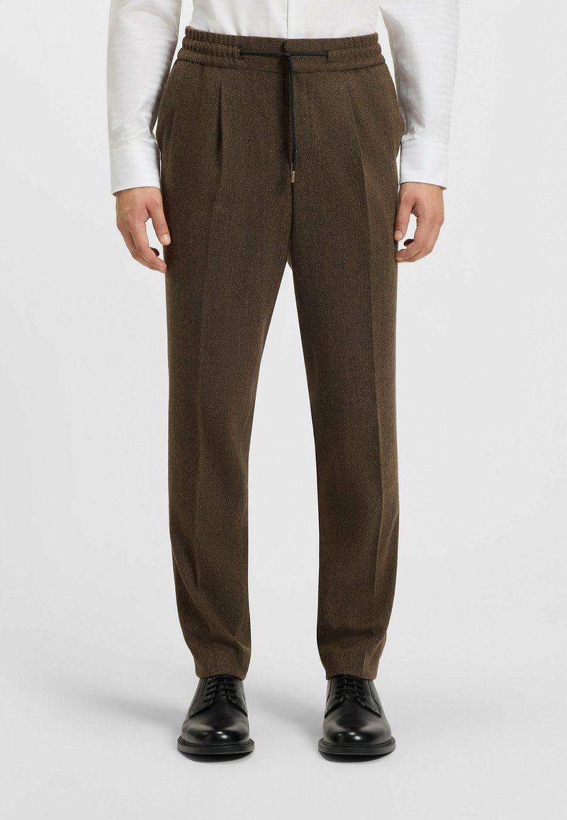 Bruine, op maat gemaakte broeken met een gestructureerd, hennepdesign. Voorzien van een elastische tailleband met een trekkoord en gestyled met zwarte schoenen.
