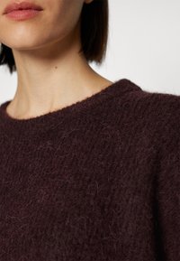 Gros plan sur une personne portant un pull texturé moelleux marron foncé avec un col rond, montrant le bas du visage et le cou.