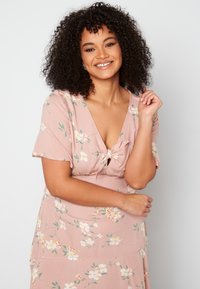 Kwiatowa sukienka z jasnoróżowego materiału, z dekoltem w serek, krótkimi rękawami i detalem w postaci węzła z przodu. Małe wzory żółtych i białych kwiatów są rozmieszczone na całej powierzchni.