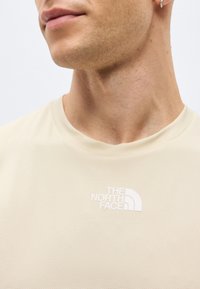 Κοντινό πλάνο ενός ατόμου που φοράει μια μπεζ μπλούζα The North Face, δείχνοντας το ντεκολτέ και το λογότυπο της μάρκας στο στήθος.