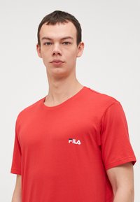 Červené bavlnené tričko s okrúhlym výstrihom a krátkymi rukávmi. Výrazné biele logo "FILA" na ľavej strane hrude. Hladká textúra.