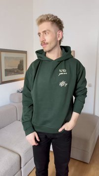 Dunkelgrüne Kapuzenjacke in lässigem Schnitt, mit weißen Text- und grafischen Details auf der Vorderseite. Schwarze Hose und strukturierter beiger Couch im Hintergrund.