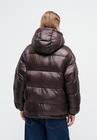 Chaqueta acolchada marrón con capucha, acabado brillante, diseño acolchado y puños elásticos, combinada con vaqueros oscuros. Vista trasera mostrando detalles de costura.