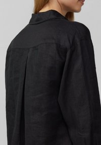 Camisa de lino negra con una textura suave, un cuello con solapa y un detalle de pliegue en la parte posterior para facilitar el movimiento.