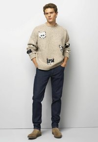 Pull beige en tricot avec motifs de chats noirs, associé à un jean bleu foncé et des bottines beiges. La texture semble douce et confortable.