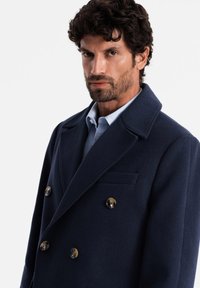 Cappotto di lana blu marina con chiusura doppiopetto, grandi bottoni neri, colletto a punta e tasche laterali; texture liscia.