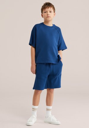 Berniukas stovintis su rankomis kišenėse, dėvintis mėlyną laisvo stiliaus marškinėlį, derančius šortus, baltas kojines su juostelėmis ir baltus sportinius batelius, neutraliame fone.