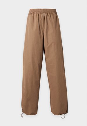 Persoon draagt een gestreepte trui met lange mouwen en losse bruine wijde broek, staat met handen in de zakken op een effen witte achtergrond.
