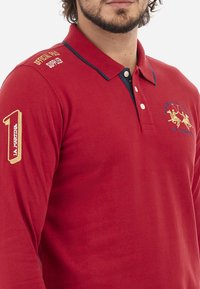Rdeča polo majica z dolgimi rokavi in zložljivim ovratnikom, mornarsko modri detajli, izšit logotip in okrasno šivanje na rokavu.