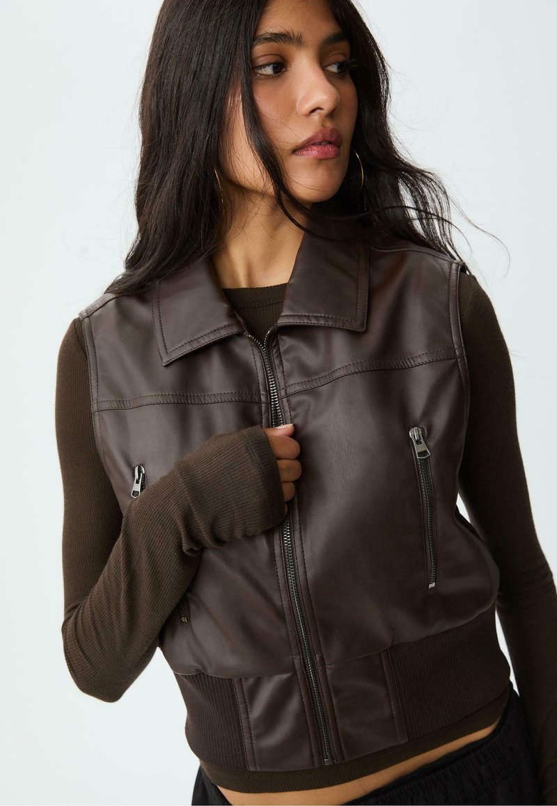Donna con lunghi capelli scuri che indossa un gilet in pelle zip-up marrone scuro sopra una maglia a maniche lunghe a costine, che guarda di lato con la mano vicina alla cerniera.