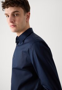 Jūras karājas zila garām piedurknēm krekls ar norādītu apkakli, pogu izgriezumu un smalku logo izšuvumu uz krūšu daļas. Gluda auduma ar pielāgotu piegriezumu.