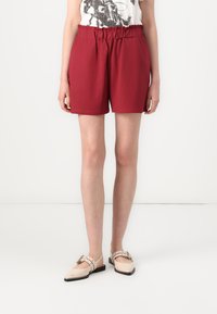 Shorts rouges à taille élastique fabriqués dans un tissu doux, avec une coupe décontractée et des poches latérales, associés à des chaussures plates à boucles de couleur claire.