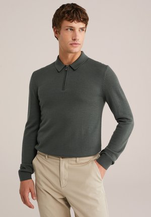 Donkergroene, lange mouwen polo trui met een halve ritssluiting kraag. Zachte textuur, aansluitend ontwerp en geribbelde manchetten. Gecombineerd met beige broeken.