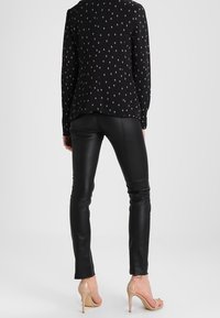 Svart långärmad blus med diamantmönster, kombinerad med figursydda svarta läderbyxor och nakna högklackade skor, vilket visar upp en slank silhuett.