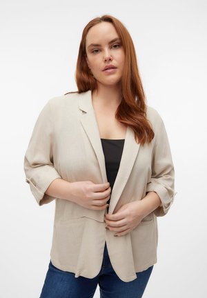 Femme aux longs cheveux roux portant un blazer beige par-dessus un haut noir et un jean bleu, ajustant le devant du blazer.