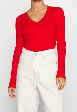 Kobieta w czerwonej bluzce z długim rękawem, z koronkowym dekoltem w kształcie litery V, oraz w jasnobeżowych jeansach z wysokim stanem, stojąca na tle jednolitego tła.