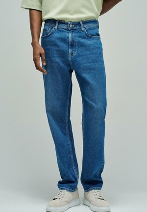 Hombre vistiendo vaqueros de corte holgado azul, zapatillas deportivas beige y una camisa de manga corta de color claro contra un fondo liso.