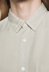 Gros plan sur une chemise beige clair à col avec trois boutons blancs fermés, montrant le cou et le haut de la poitrine.