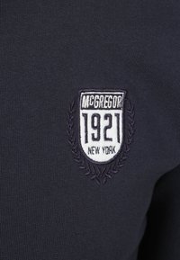 Marine gebreid kledingstuk met een wit geborduurd embleem met "MCGREGOR 1921 NEW YORK" en laurieraccenten. Zachte textuur, fitted vorm.