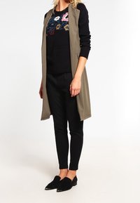 Longue veste vert olive portée sur un pull noir en tricot avec broderies florales colorées, associée à un pantalon noir slim et des chaussures noires à enfiler.