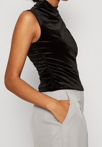 Femme portant un haut noir sans manches froncé et un pantalon gris clair, avec sa main droite dans la poche, devant un fond uni.