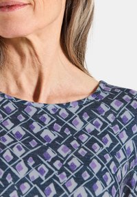Bluse mit blauem und lila Muster, das ein geometrisches Design aus Rauten und Quadraten zeigt, mit rundem Ausschnitt und kurzen Ärmeln.