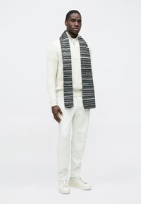 Pull blanc en maille avec col à zip, pantalon clair et écharpe grise rayée. Les baskets sont beiges. Fond neutre.