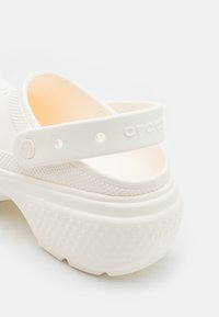 Crocs bianchi con un design testurizzato, dotati di una cinghia posteriore e punteggiature rialzate sulla suola. Materiale liscio con una leggera lucentezza e bordi arrotondati.