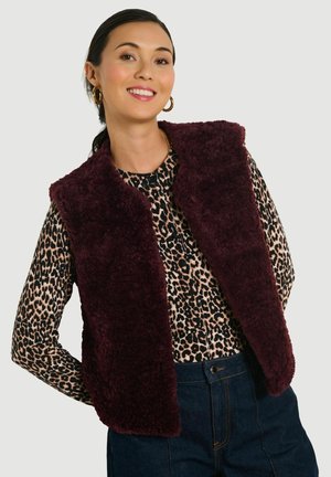 Lachende vrouw met donker haar, die gouden ronde oorbellen draagt, een bordeaux faux fur-vest, een blouse met luipaardprint en donkere blauwe spijkerbroek.