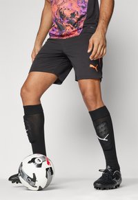 Μαύρες αθλητικές βερμούδες με πορτοκαλί λογότυπο, τοπ με μοτίβο σε μωβ και πορτοκαλί, μαύρες κάλτσες ως το γόνατο και μαύρα ποδοσφαιρικά παπούτσια δίπλα σε μια μπαλάρα ποδοσφαίρου με μοτίβο.