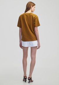 Kurzärmliges, senfgelbes Oberteil aus glattem Stoff, kombiniert mit weißen Shorts. Das Design zeichnet sich durch eine lockere Passform und einen runden Ausschnitt aus.
