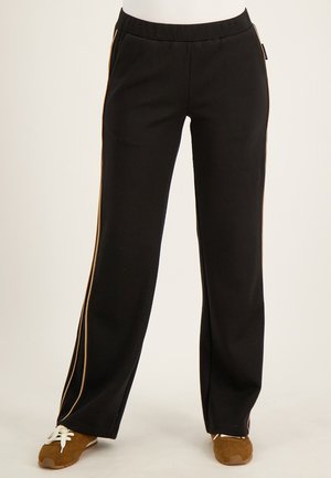 Zwarte wijde broek met beige zijstrepen gedragen met bruine veterschoenen op een neutrale achtergrond.