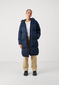 Tumši zila pūkaino mēteli ar kapuci, ar quilted dizainu un priekšējo rāvējslēdzēju. Saskaņots ar bēšiem biksēm un tumšiem sporta apaviem.