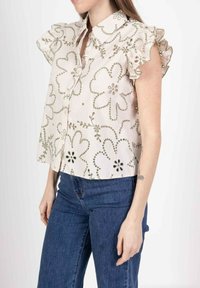 Blouse à manches courtes et col en beige clair avec broderies florales, manches volantes et boutons décoratifs. Portée avec un jean en denim bleu.