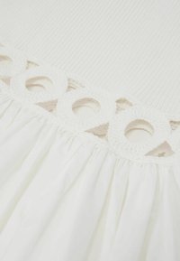 Weißer Stoff mit gerippter Struktur oben und gesammeltem glattem Material unten, getrennt durch ein Band aus gehäkelter runder Spitze.
