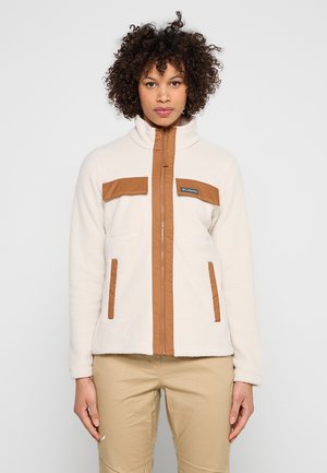 Mujer con chaqueta de forro polar crema con adornos marrones y cremallera, de pie contra un fondo liso gris claro, mirando hacia adelante.