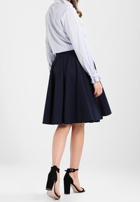 Chemise à rayures bleues et blanches à boutons, associée à une jupe patineuse navy foncé. Des sandales à talons noirs ajoutent du contraste. Textures lisses, design classique.