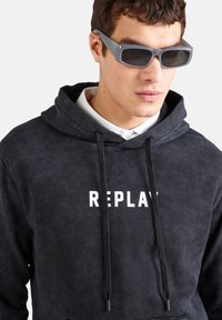 Černá mikina s texturovaným povrchem, s bílým logem "REPLAY", šňůrkami a s bílou košilí s límečkem pod ní. Šedé sluneční brýle.