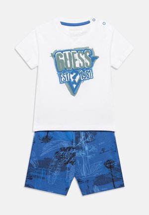 Biała koszula z krótkim rękawem z niebieskim logo „GUESS EST 1981” oraz niebieskie szorty z abstrakcyjnym wzorem tropikalnych roślin.