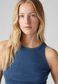 Débardeur bleu à côtes avec un col haut et de larges emmanchures. Le tissu a une texture douce, offrant un look ajusté.