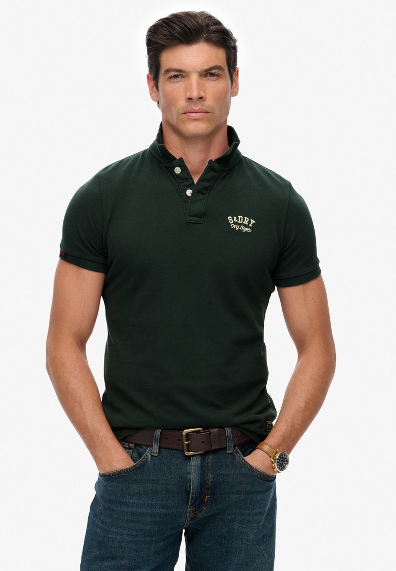 Donkergroen poloshirt met een kraag met knopen, geborduurde merknaam en een gestructureerde stof. Gecombineerd met een blauwe spijkerbroek en een bruine leren riem.