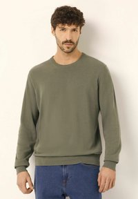Pulover verde măsliniu cu guler rotund, mâneci lungi, material texturat, cu manșete și tiv nervurat, combinat cu blugi denim albaștri.