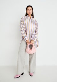 Chemise rayée en lavande, jaune et blanc, associée à un pantalon large gris ; tenant un sac en tissu rose et portant des bottines à talons blocs roses.