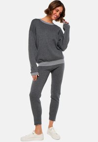 Une femme souriante dans un pull en tricot gris foncé et un pantalon ajusté assorti, posant avec une main près du visage, portant des baskets blanches sur un fond blanc.