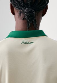 Kremlam brīvs džersijs ar zaļu apkakli, kas izgatavots no strukturēta, elpojoša auduma, un aizmugurē ir zaļā krāsā izšūta "KidSuper" logo.